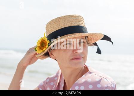 Woman in a pink dress with white polka dots and a boater with a sunflower, thoughtfully looking away,on a blurred sea background. Stock Photo