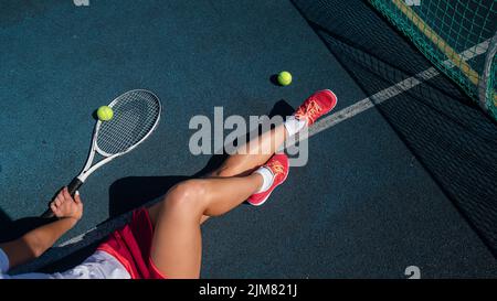 A faceless girl in a sports skirt sits on a tennis court and holds a rocket Stock Photo