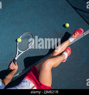 A faceless girl in a sports skirt sits on a tennis court and holds a rocket. Top view of female legs. Stock Photo