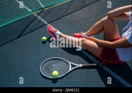 A faceless girl in a sports skirt sits on a tennis court and holds a rocket. Top view of female legs. Stock Photo
