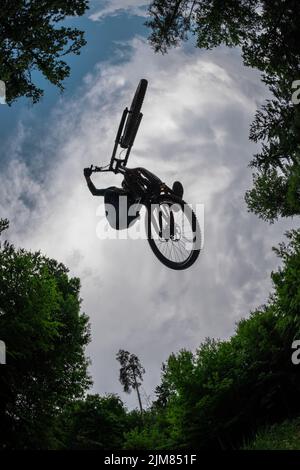 Silhouette of a mountain biker jumping over camera and performing tail whip. Extreme photo of mtb racer jumping surrounded by trees and cloudy sky. Go Stock Photo