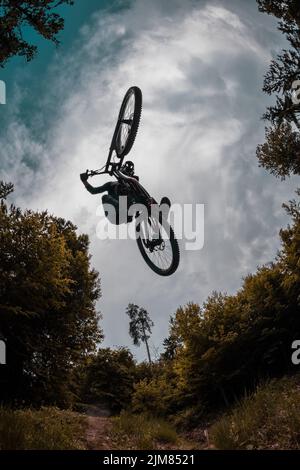 Silhouette of a mountain biker jumping over camera and performing tail whip. Extreme photo of mtb racer jumping surrounded by trees and cloudy sky. Go Stock Photo