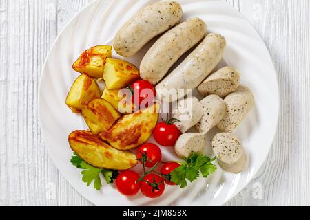 Weisswurst, bavarian white sausage of minced veal, pork back bacon, spices and parsley on white plate with roast potatoes, fresh tomatoes, horizontal Stock Photo