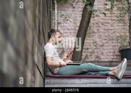 Focused young man entrepreneur sitting outside with laptop and smartphone, enjoy open-air workday Stock Photo