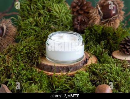 Opened glass jar full of cream on wooden piece between green moss, pine cones and nuts, closeup, mockup. Skincare beauty product package. Natural Orga Stock Photo