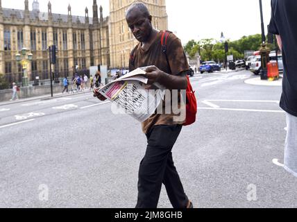 London, England, UK. Black man reading a newspaper (Evening Standard: 15th July 2022) in Westminster, walking past the Houses of Parliament Stock Photo