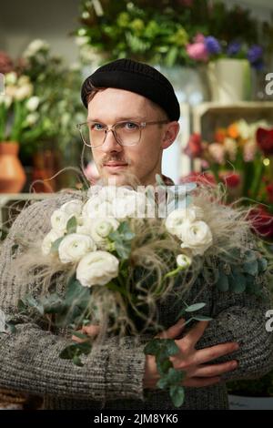 Good looking male florist wearing sweater, beanie and eyeglasses holding flower bouquet. Small business concept. Bearded man with bouquet of white roses in flower shop. High quality vertical image Stock Photo