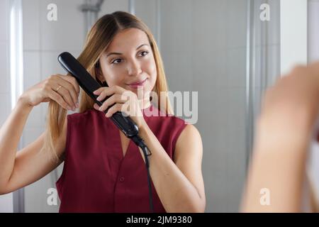 Beautiful young woman using steam straightener to style hair at the mirror on bathroom Stock Photo