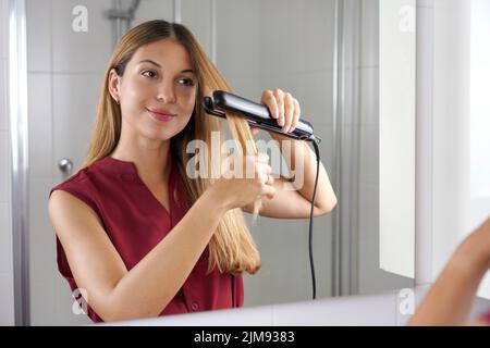 Hispanic young woman using steam straightener to style hair in the mirror on bathroom Stock Photo