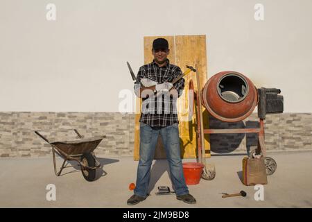 Image of a smiling construction worker with folded arms at work on a construction site with a wheelbarrow and concrete mixer in the background. Diy Stock Photo
