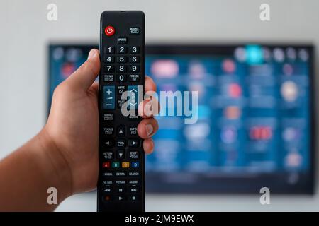 Smart tv and hand pressing remote control. Hand holding TV remote control with a television in the background. Close up. Stock Photo