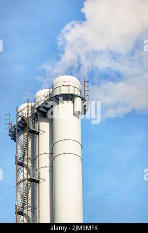 A fragment of pipes of a heating plant emitting white smoke into the blue sky on a bright day. Closeup, copy space, vertical image. Stock Photo