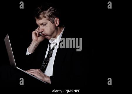 Stressed mature man listening on cell phone while working on laptop in the darkness Stock Photo