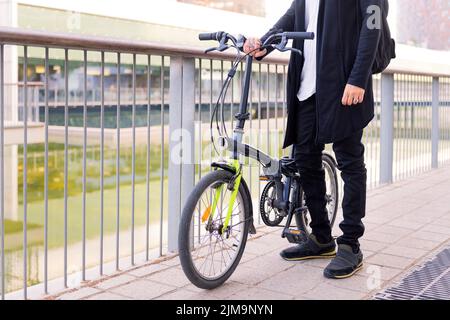 unrecognizable young man wearing trendy clothes and holding his folding bike in a city park, concept of urban lifestyle and sustainable mobility, copy Stock Photo