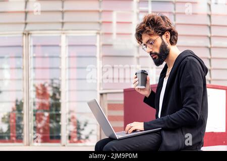 stylish young man working with a laptop computer drinking coffee in front of an office building, concept of technology and urban lifestyle, copy space Stock Photo