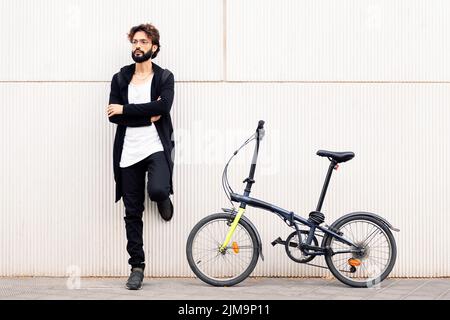 stylish young man with beard leaning against a white wall next to his folding bike, concept of urban lifestyle and sustainable mobility, copy space fo Stock Photo