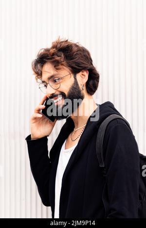 portrait of a stylish young man with beard walking in front of a white wall talking on mobile phone, concept of urban lifestyle and technology of comm Stock Photo