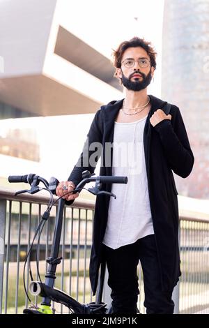 young caucasian man with beard wearing trendy clothes and walking with his folding bike through the city, concept of urban lifestyle and sustainable m Stock Photo