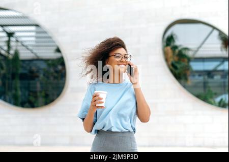 Phone talk, communication. Positive young african american curly woman, in formal stylish wear, with glasses, walking outdoors, holding takeaway coffee cup, talking by smartphone, smiling happily Stock Photo