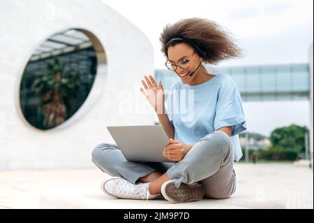 Busy successful african american young woman with headset, business lady, sitting outdoors, holds online video conference with colleagues by laptop, records online video tutorial, smiling Stock Photo