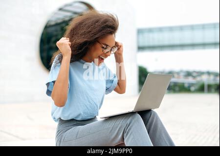 Emotional cheerful african american young woman with glasses, uses laptop while sitting outdoors, gesturing hands, with excitement looks at screen, read unexpected news, celebrating success, smiles Stock Photo