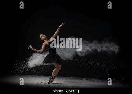 An emotional Southeast Asian ballet dancer in a black dress with powder performing a move on a black background Stock Photo