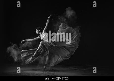 A grayscale shot of an emotional Southeast Asian ballet dancer in a dress with powder performing a move on a black background Stock Photo