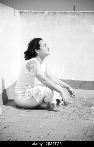 A black and white photo of a young woman in a white suit sunbathing alone on the terrace. Stock Photo