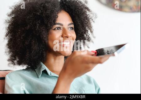 Smiling African-american woman with curly hair holding phone and speaking on speakerphone, multiracial girl recording and sending voice message, using voice typing feature on smartphone at home Stock Photo