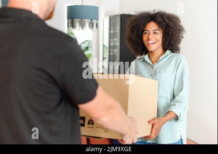 Back view of courier giving cardboard box to happy african american woman. Cheerful multiracial girl with natural Afro hairstyle receiving a package at home from a delivery guy, ordered online Stock Photo