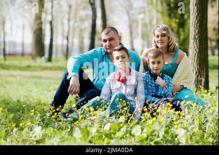 Happy pregnant family with two sons, dressed in a turquoise clothes sitting on grass with flowers at park Stock Photo