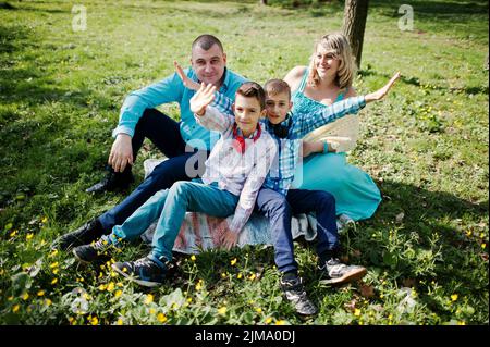 Happy pregnant family with two sons, dressed in a turquoise clothes sitting on grass with flowers at park Stock Photo