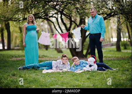 Happy pregnant couple with two sons holding girls' baby clothes on string  background spring nature Stock Photo