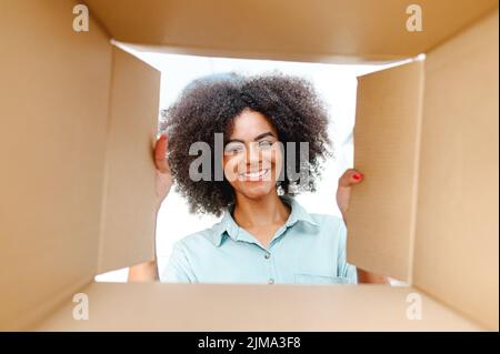 POV view from inside box at satisfied biracial young woman with an Afro hairstyle unpacking package box, excited happy African woman looks inside delivery box. Glad customer of e-shop Stock Photo
