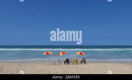 scenic view of red and yellow beach umbrellas with plastic chairs in a row on sand at the beach against clear blue sky and space for text Stock Photo