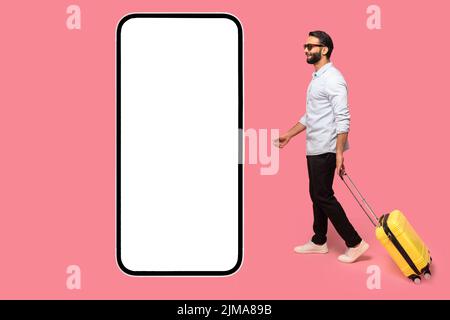 Mobile app for travelers. Indian man in sunglasses with yellow luggage suitcase walking to huge smartphone with empty display, copy space, mockup Stock Photo
