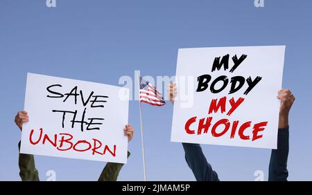 For and against legal abortion. Two women holdig signs saying Save The Unborn and My Body My Choice. American flag and blue sky background with copy s Stock Photo