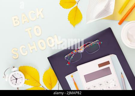 Back to school concept. School supplies on a tablet with glasses on a blue background with yellow, autumn leaves, alarm clock and wooden inscription B Stock Photo
