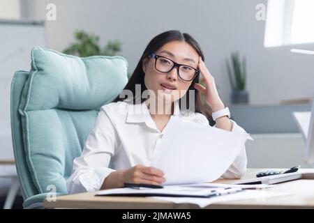 Young beautiful Asian woman in glasses and business clothes holding her head with her hand, sitting in a chair at a table in the office, holding documents in her hands Stock Photo