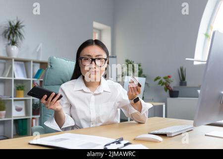 Fraud. Upset young beautiful Asian woman holding phone and credit card. A disappointed young lady lost her life savings to a scam. Sitting at the desk in the office Stock Photo