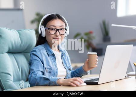 Online training. Young beautiful Asian student girl in glasses studies at a laptop in headphones, drinks coffee. Learns remotely, sits at a desk in a modern office, looks at the camera. Stock Photo