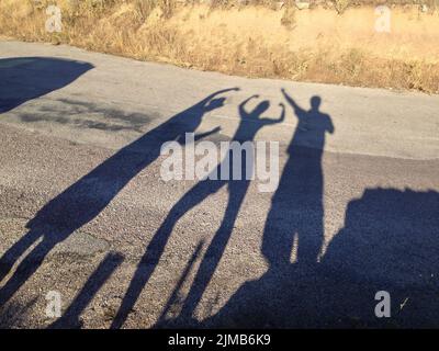 Long shadows of three people in different poses with their arms in the air on a road Stock Photo