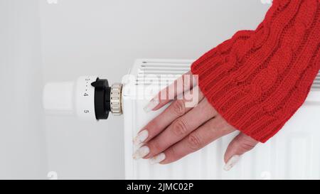 Close-up of a woman in red sweater warms her hands over the battery on background of white wall.Macro photo of woman's hands,copy space.Concept of sav Stock Photo