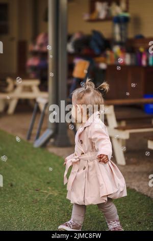 Toddler girl with pigtails at day care center wearing cute pink coat surrounded by bubbles, no face Stock Photo