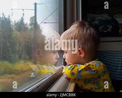 A bright, cute three-year-old boy riding a train looks out the window, behind which a dull landscape sweeps through. Stock Photo