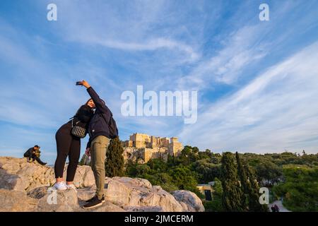 A couple are taking a selfie on a hill across the Acropolis rock with the Acropolis in the background on a sunny afternoon. Stock Photo