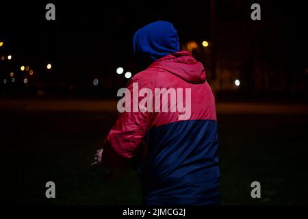 Guy in dark in hood. Unknown person on street. Man in red jacket hides his face. Modern style of clothing. Stock Photo