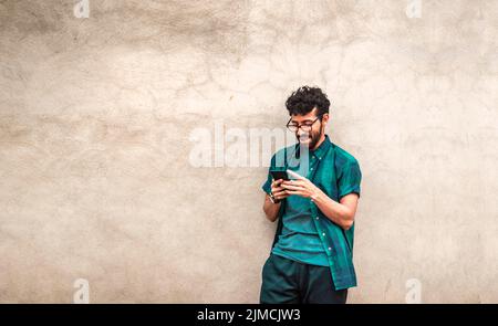 Happy handsome guy leaning against the wall using his cell phone, happy latin man with his cell phone leaning against a wall with copy space, Person Stock Photo