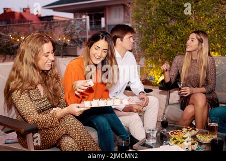 Group of smiling friends chatting and having dinner with tasty appetizers while sitting at table on terrace in evening time Stock Photo