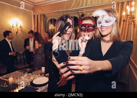 Glad female friends in masquerade masks taking self portrait on smartphone while having dinner together in modern restaurant with friends Stock Photo